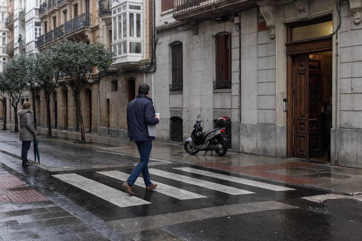 Un agente inmobiliario cruza un paso de cebra mojado en Indautxu, Bilbao, a primera hora de la mañana, con una carpeta bajo el brazo y un café, hacia el portal abierto de un edificio con miradores blancos.