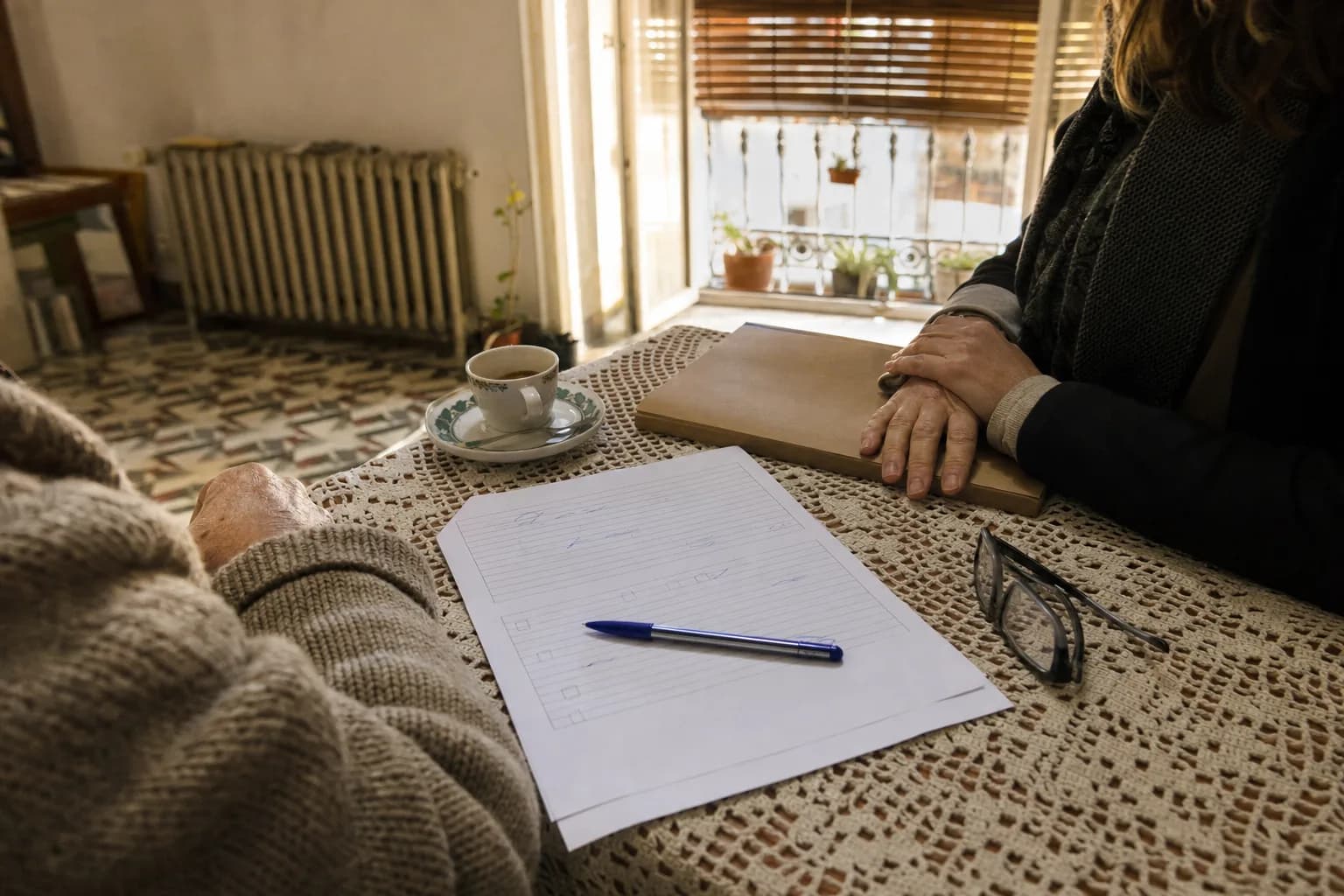 Una ficha de captación en papel, a medio rellenar y con un bolígrafo encima, sobre la mesa del salón de un piso del Realejo, en Granada, durante una visita de valoración; al fondo, desenfocadas, las manos de la agente sobre una carpeta y la manga de punto del propietario.