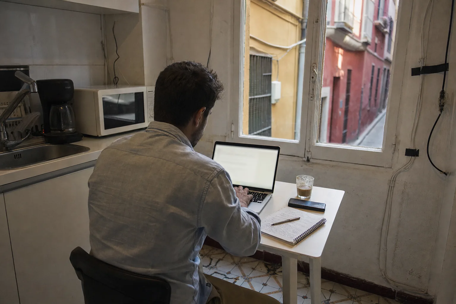 Un agente inmobiliario de espaldas escribe en un portátil sobre una mesa pequeña en la cocina de una oficina del Soho de Málaga, junto a una cafetera, un microondas y una libreta con notas a mano.