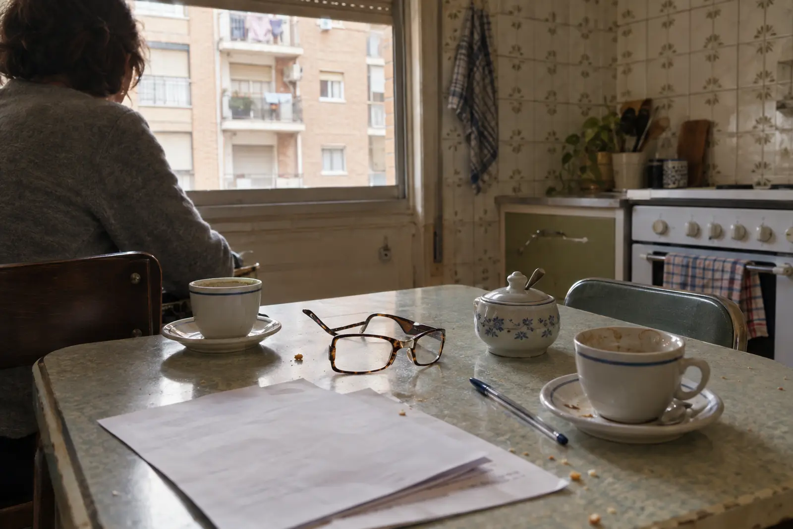 Mesa de cocina en un piso de Zaragoza a media tarde, con varias hojas grapadas, dos tazas de café y unas gafas de leer; al fondo, desenfocada, la espalda de una persona sentada.