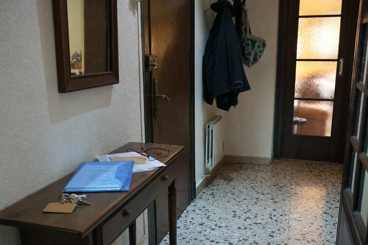 Recibidor de un piso de Murcia al anochecer: junto a la puerta, un mueble de madera con un manojo de llaves con etiqueta de cartón, una carpeta azul de plástico con papeles y unas gafas, sobre un suelo de terrazo.