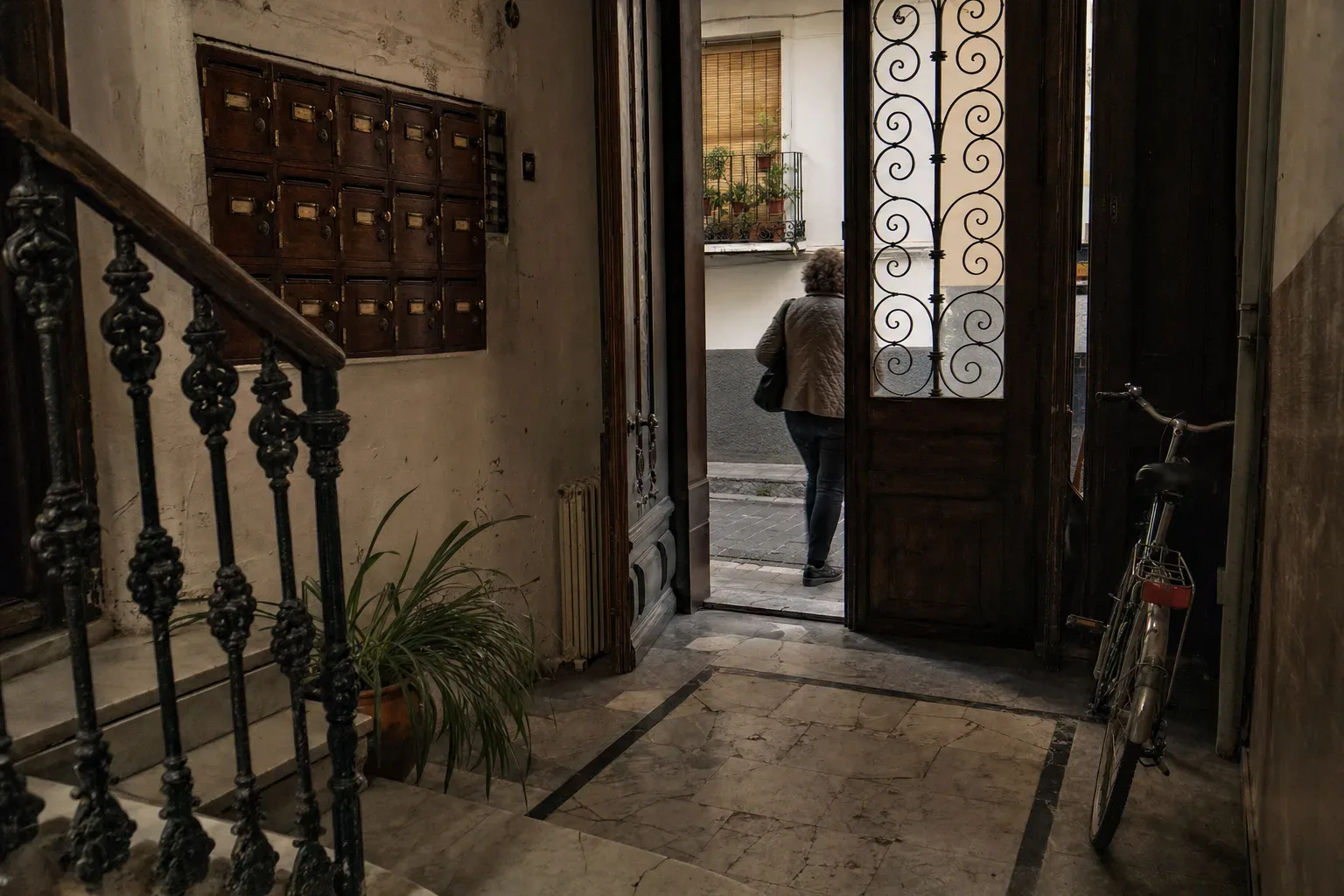Buzones antiguos de madera oscura y latón en el portal de un edificio del Realejo, en Granada, con las tarjetas de nombre en blanco, junto a una escalera con barandilla de hierro y una bicicleta apoyada en la pared.