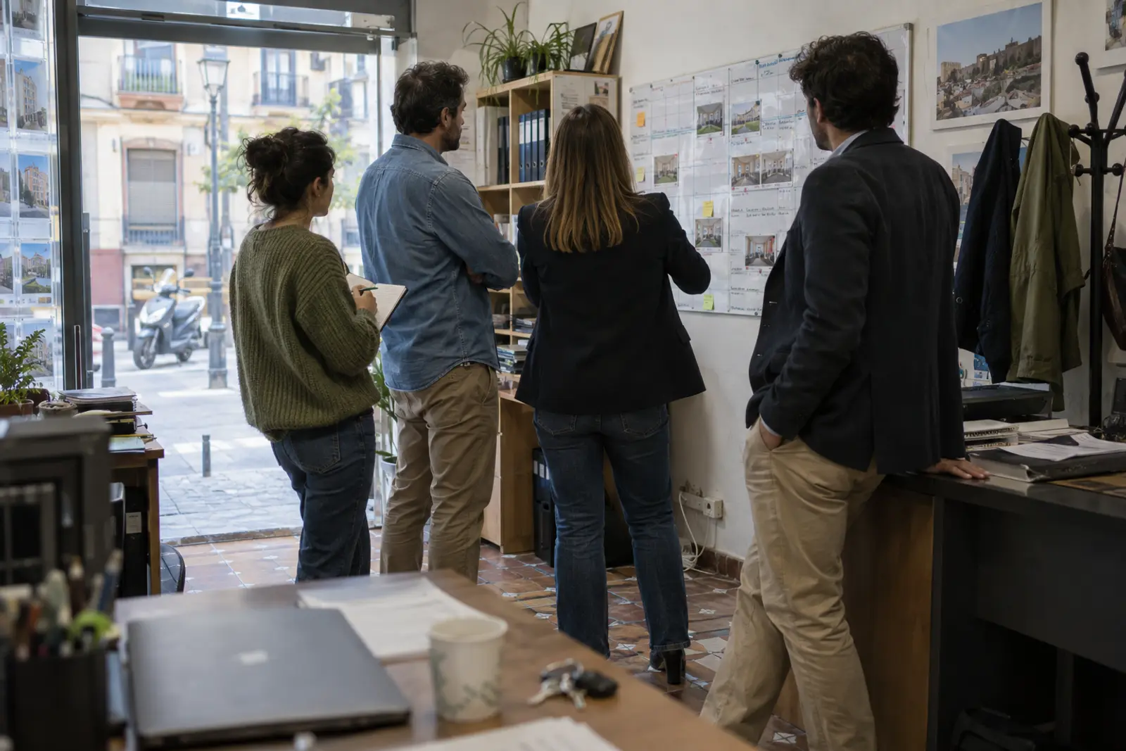 Reunión de pie de un equipo de cuatro agentes inmobiliarios en una oficina pequeña, vista desde el fondo de la sala, junto a un planificador de pared a primera hora de la mañana.