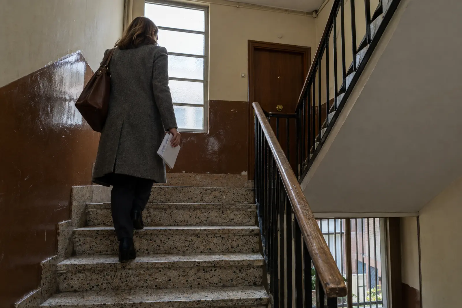 Agente inmobiliaria de espaldas subiendo la escalera de un edificio de vecinos de los años setenta en Valladolid, con barandilla de hierro y luz de media mañana entrando por la ventana del rellano.